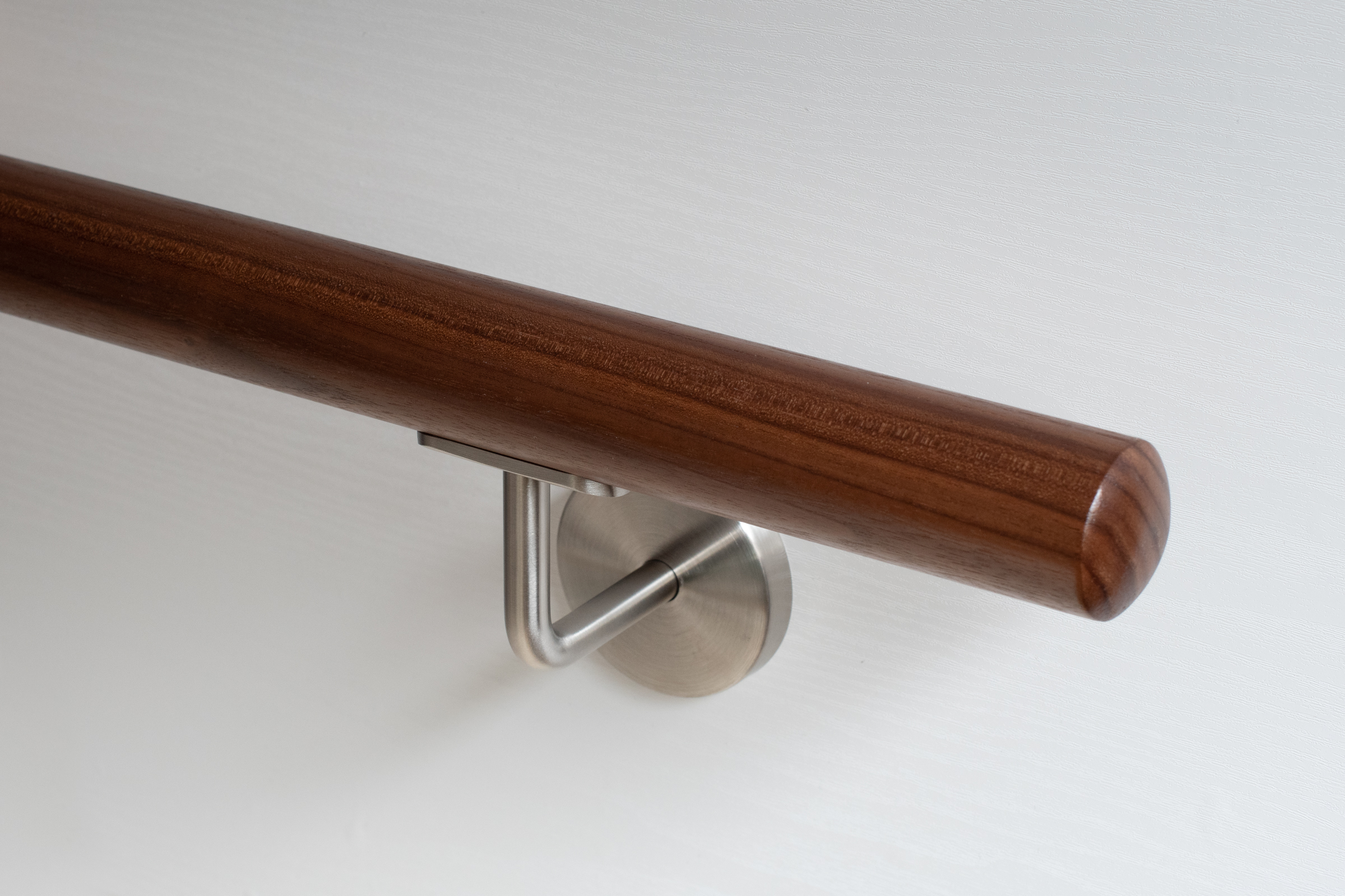Corrimano rotondo in noce con supporto a parete inox su parete bianca, legno marrone scuro, primo piano dell'estremità