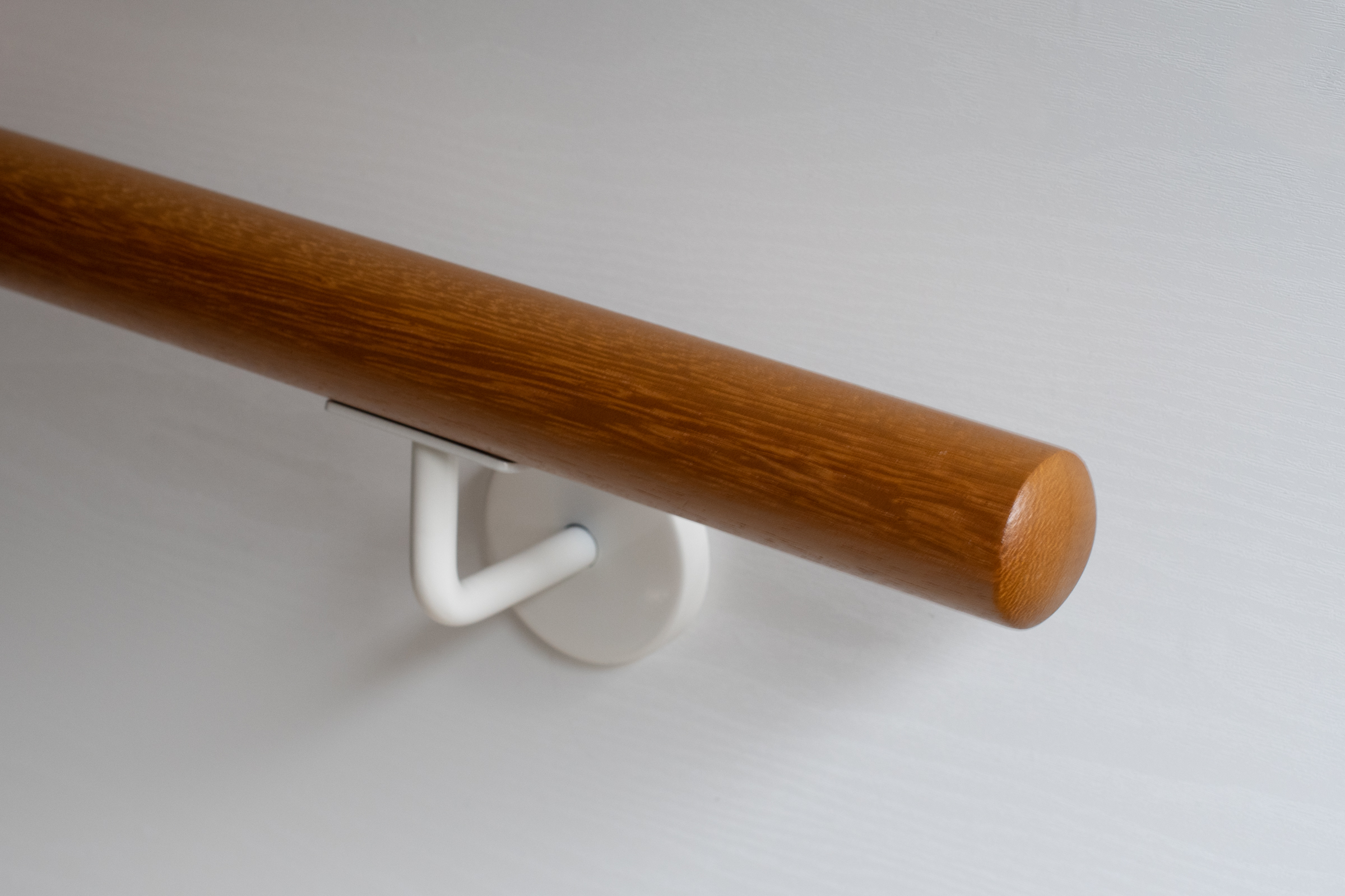 Corrimano rotondo in iroko con supporto a parete bianco su parete bianca, legno bruno dorato, primo piano dell'estremità