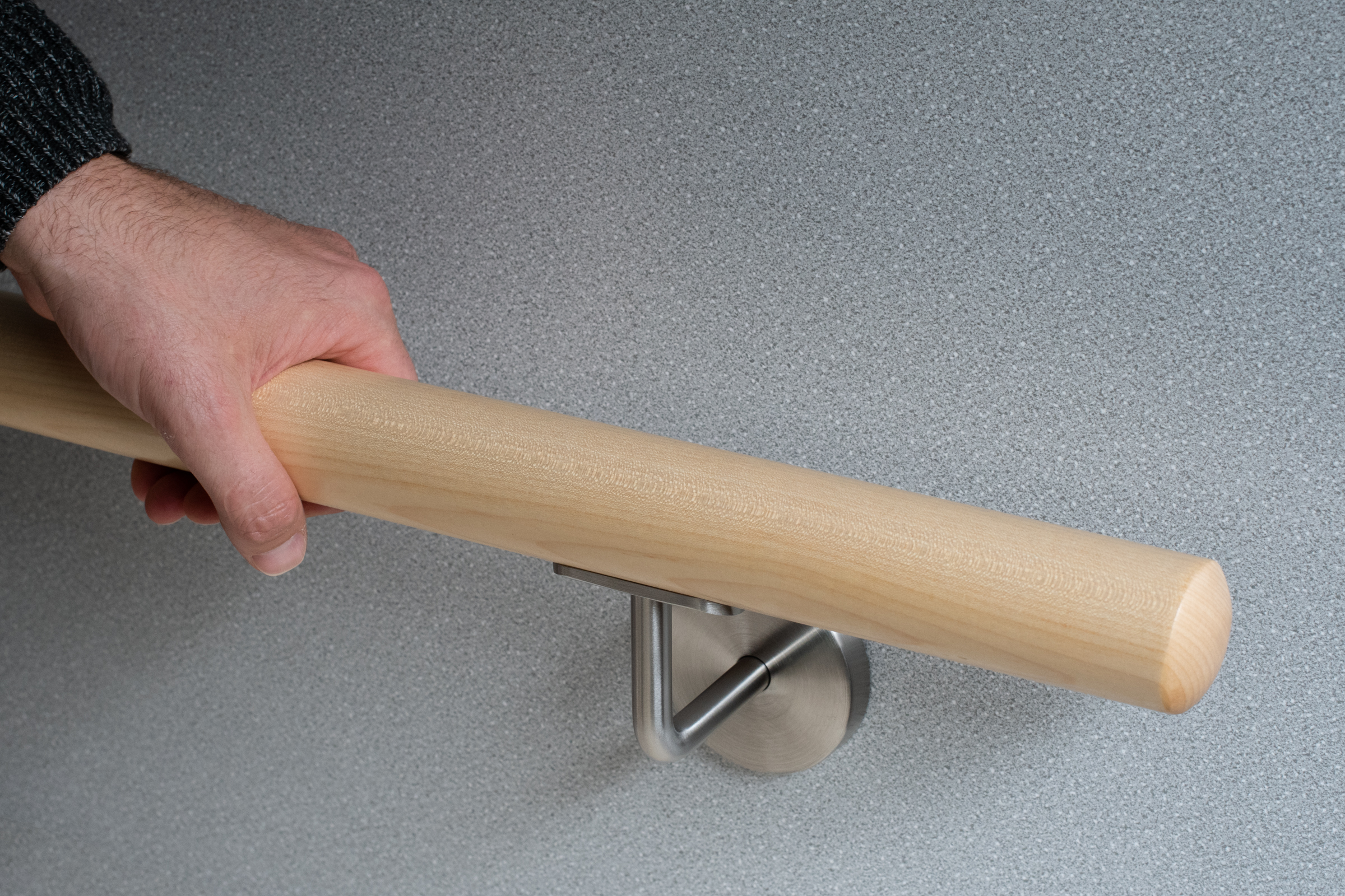 Corrimano rotondo in acero con supporto inox su parete grigia, una mano impugna il corrimano in legno chiaro, primo piano