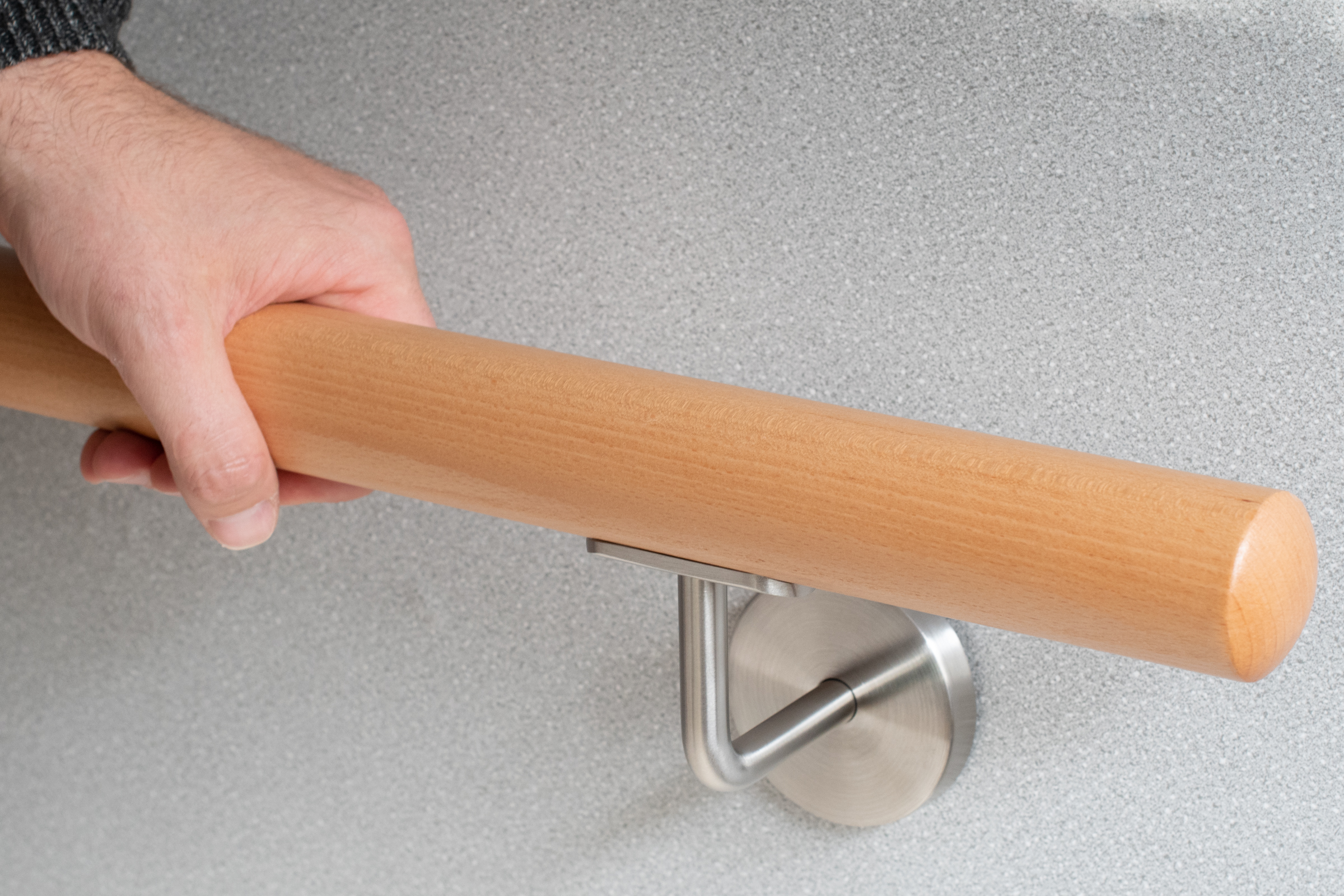 Corrimano rotondo in faggio su parete grigia con supporto inox, una mano afferra il corrimano, dettaglio della presa