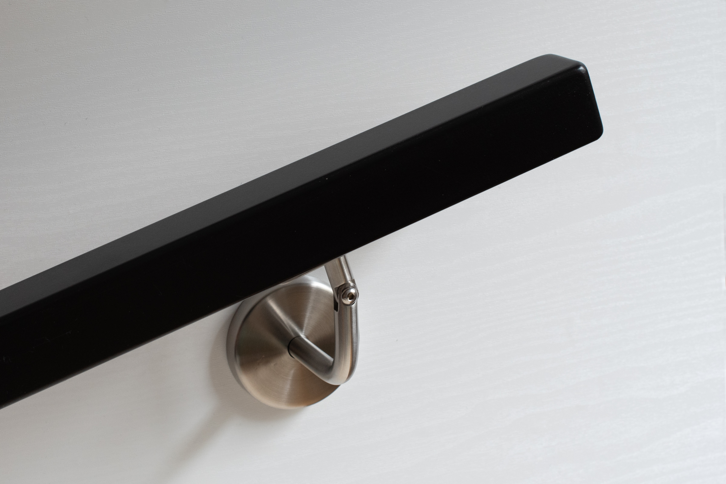 Corrimano quadrato verniciato nero con supporto a parete inox su parete bianca in legno, superficie opaca, vista obliqua