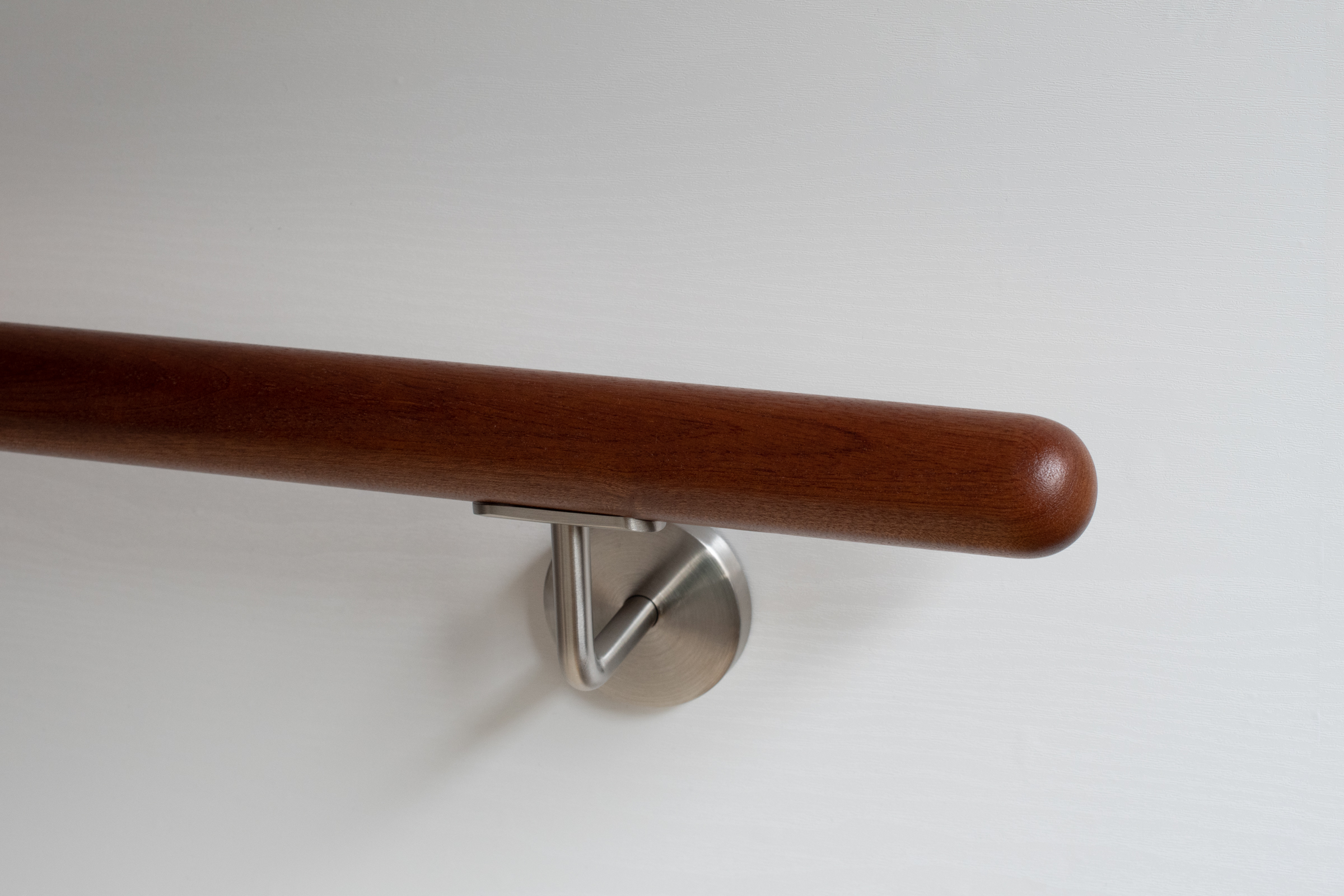 Corrimano rotondo in mogano con supporto inox su parete bianca, legno rossastro, primo piano dell'estremità arrotondata