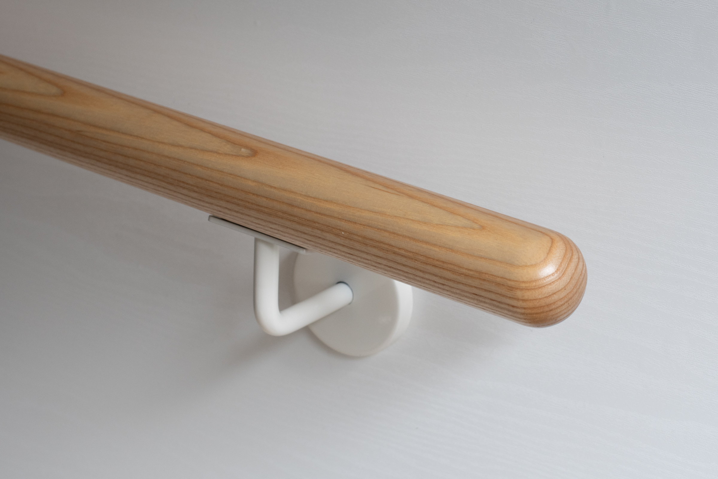 Corrimano rotondo in frassino con supporto a parete bianco su parete bianca, legno chiaro con venatura marcata, primo piano