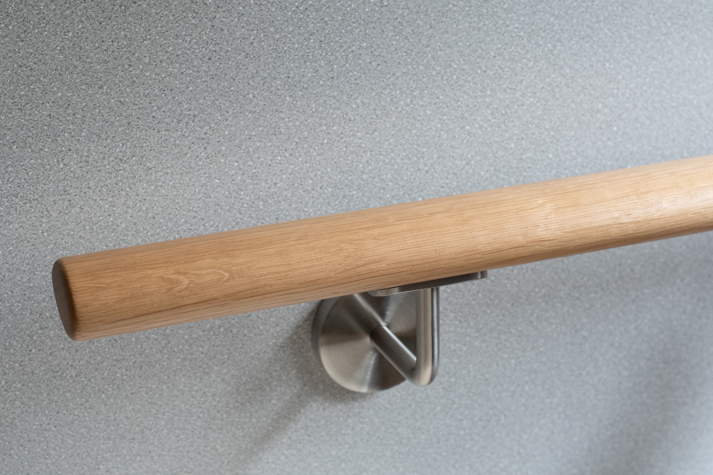 Corrimano rotondo in rovere con supporto a parete inox su parete grigio chiaro, primo piano con venatura del rovere marcata