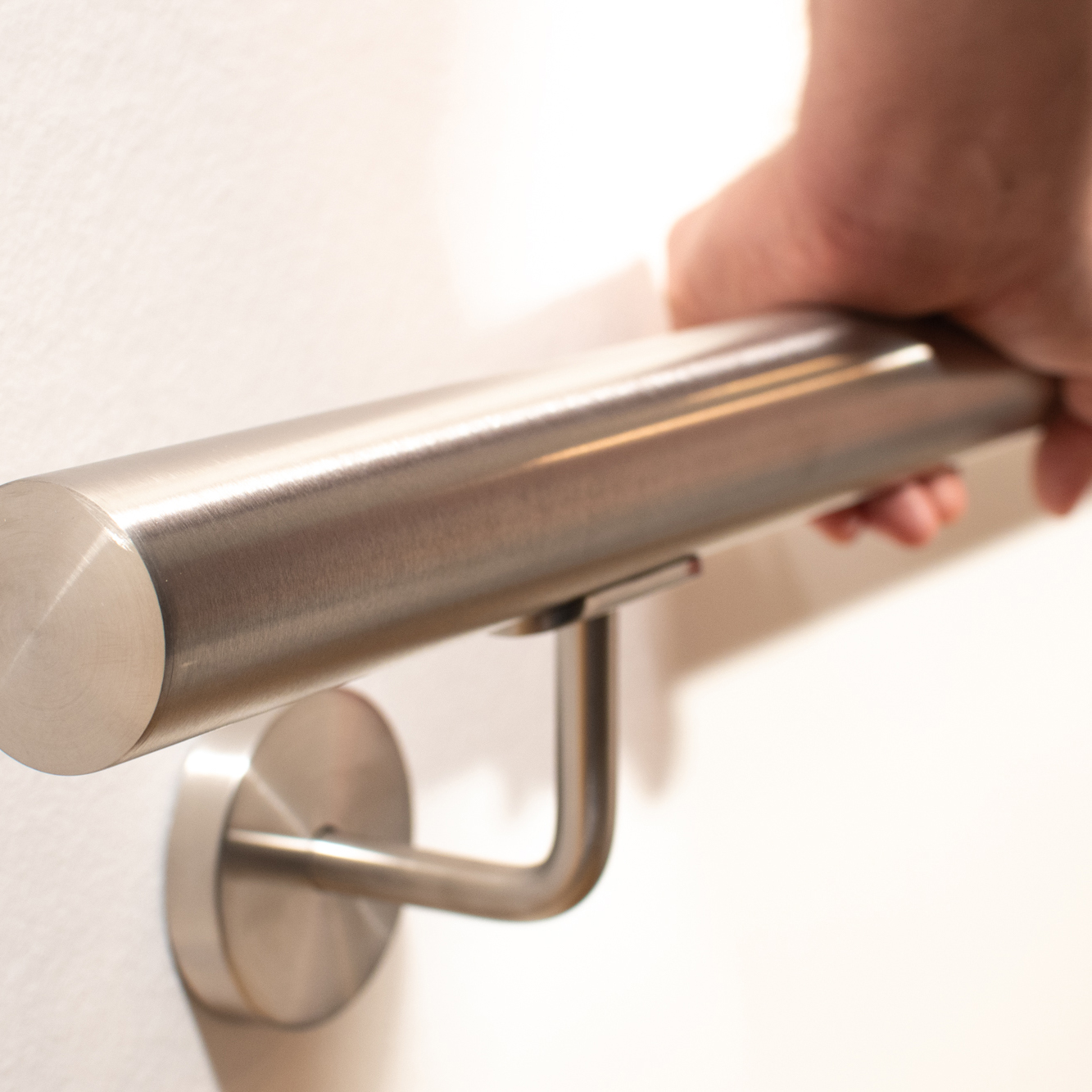 Corrimano rotondo in acciaio inox su parete bianca con supporto a parete, una mano afferra il corrimano tubolare spazzolato