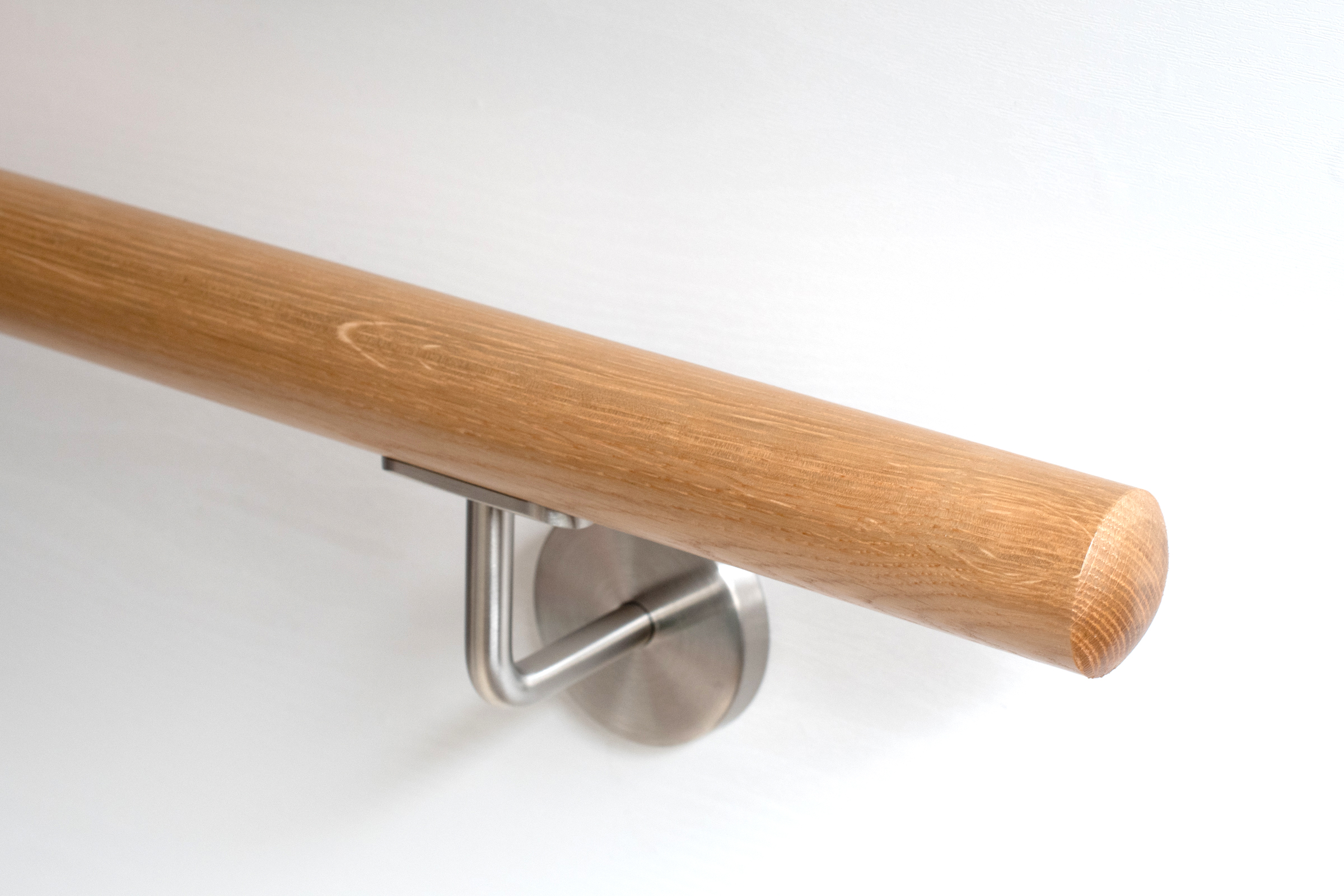 Corrimano rotondo in rovere rosso con supporto inox su parete bianca, primo piano dell'estremità arrotondata con venatura
