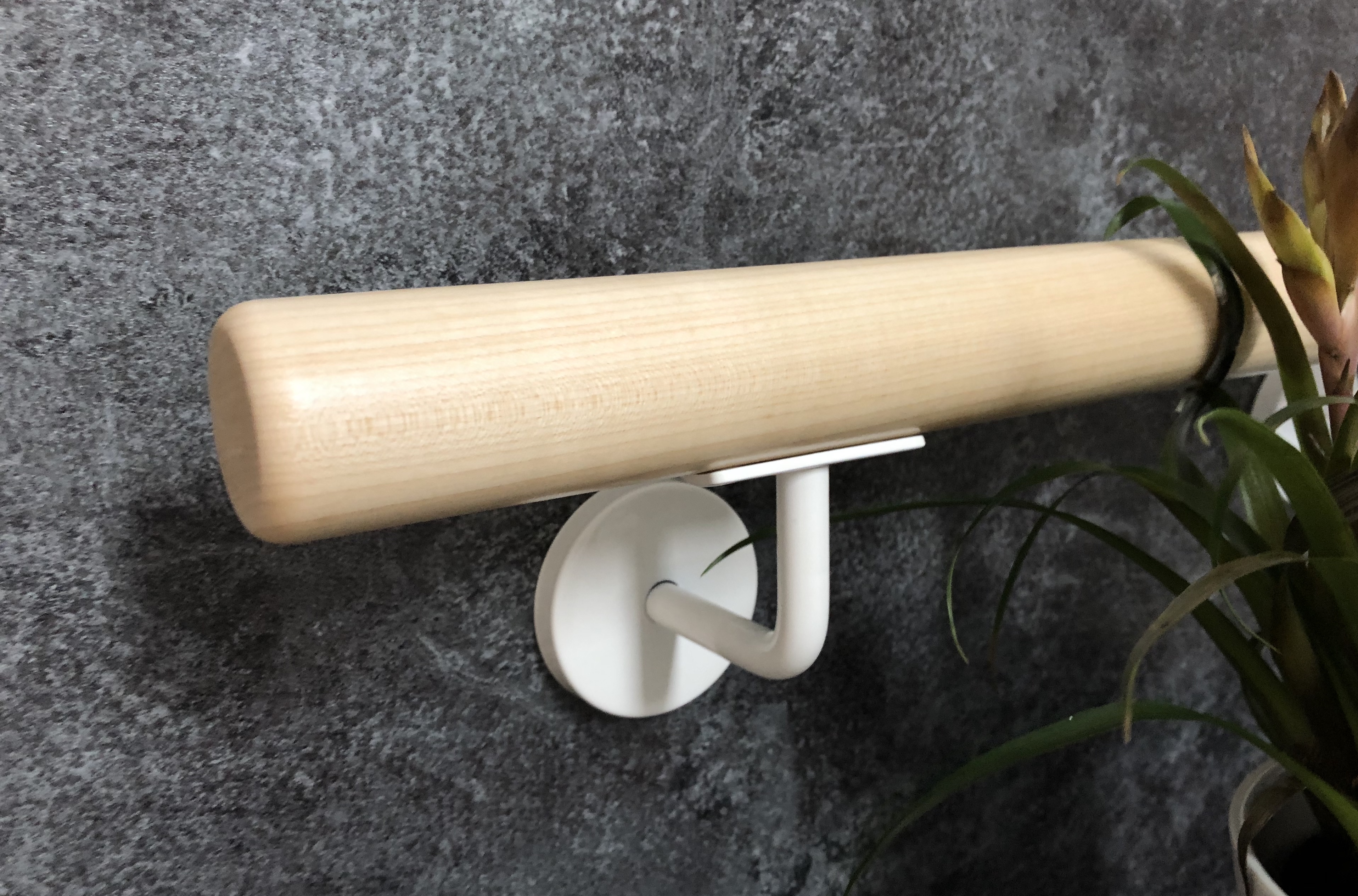 Corrimano in legno rotondo chiaro con supporto verniciato bianco su parete in pietra grigio scuro, pianta verde a lato